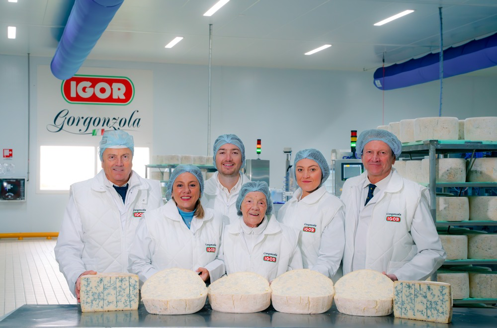 A group of six workers in white coats and hairnets at the IGOR Gorgonzola cheese factory in Novara, Italy, with cheese wheels in front of them.