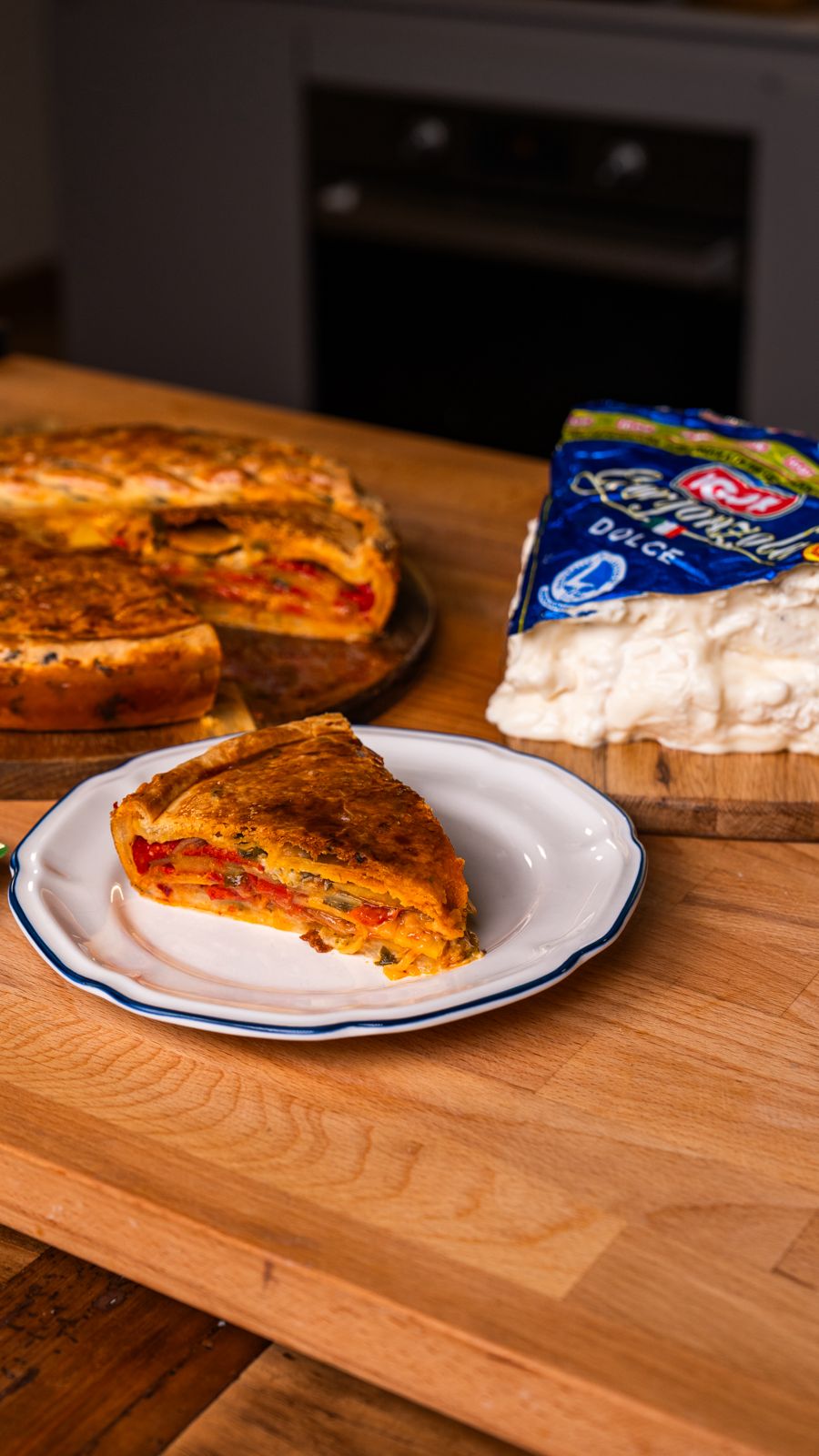 Fetta di torta salata con Gorgonzola Igor, peperoni, patate e nduja su un piatto, con formaggio Gorgonzola e altri ingredienti sul tavolo.
